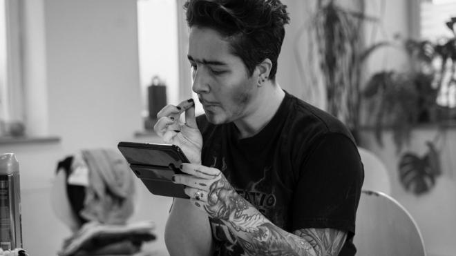 A black and white picture of a person, that is holding a pocket mirror and is using make up to paint a beard onto their face.